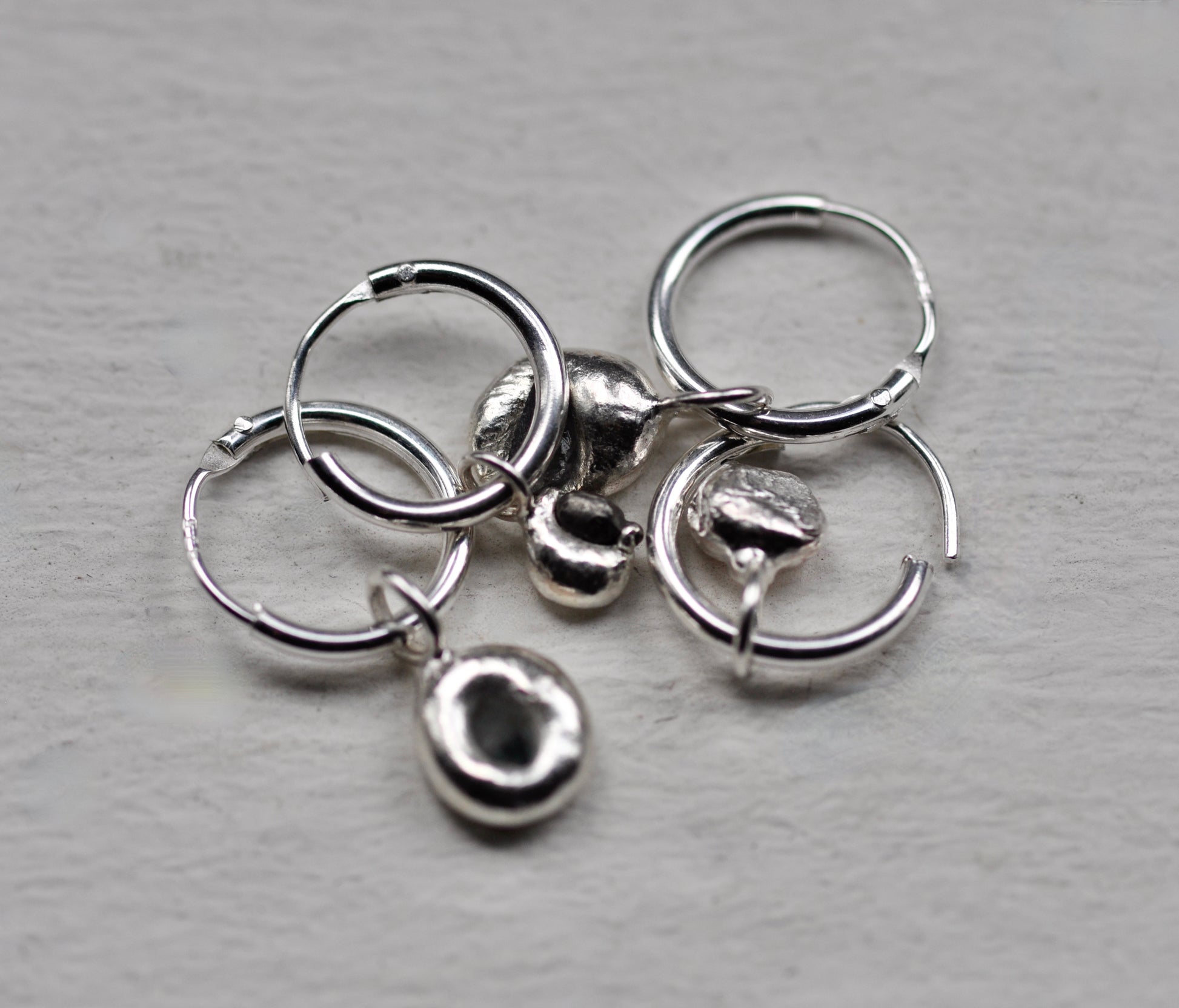 A set of sterling silver jewelry earrings, including three small hoop earrings, two textured dome charms, and two tiny textured charms, arranged on a light gray textured surface.