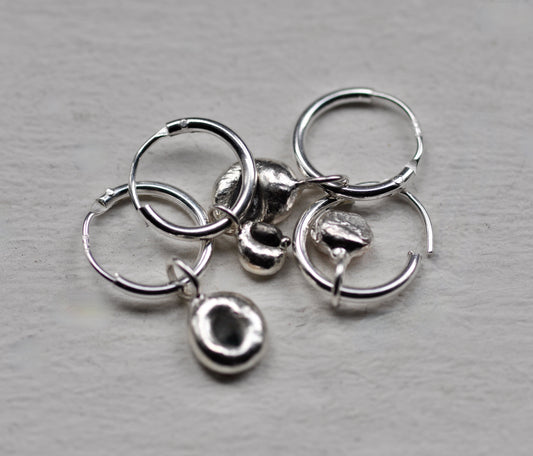 A set of sterling silver jewelry earrings, including three small hoop earrings, two textured dome charms, and two tiny textured charms, arranged on a light gray textured surface.